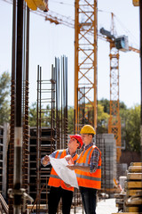 Construction manager and engineer dressed in orange work vests and hard helmets explore construction documentation on the building site near the steel frames