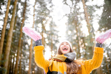 Portrait of a young exccited woman dressed in bright winter clothes enjoying snow falling in pine forest