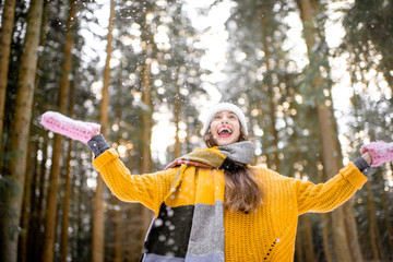 Portrait of a young exccited woman dressed in bright winter clothes enjoying snow falling in pine forest