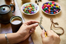 Close-up Of Woman Sitting At The Table Drinking Coffee And Making Handmade Accessories From Colorful Beads