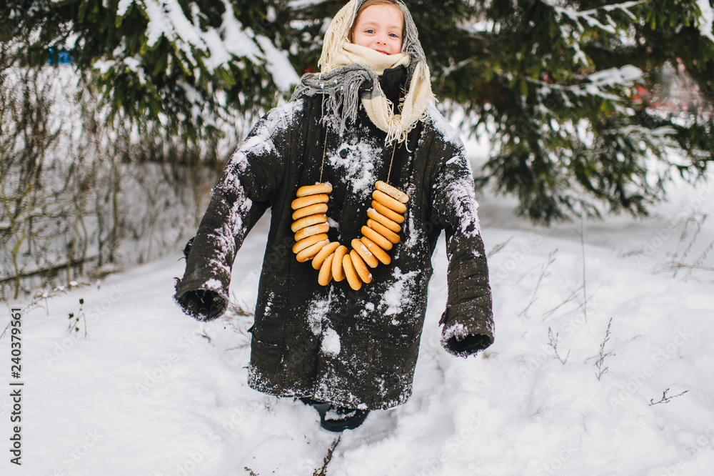 Little funny girl in big adult oversized clothes posing on snow in winter  day. Lovely female child with bunch of bagels looking at camera outdoor.  Russian cultural traditional holiday celebrating.. Stock Photo |