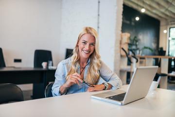 Wall Mural - Beautiful blonde woman at workplace. Holding eyeglasses and looking at camera.