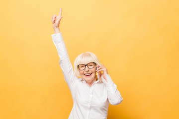 Portrait of a cheerful senior woman gesturing victory isolated over yellow background. Using phone.