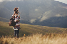 Stylish Hipster Girl In Hat Walking On Top Of Mountains. Happy Young Woman With Backpack Exploring Sunny Mountains. Travel And Wanderlust Concept. Amazing Atmospheric Moment