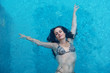 schöne reife Frau im besten Alter mit dunklen Locken im Bikini schwimmt mit erhobenen Armen schwerelos elegant glücklich im türkis blauen Wasser im Pool