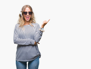 Poster - Beautiful young blonde woman wearing headphones and sunglasses over isolated background with a big smile on face, pointing with hand and finger to the side looking at the camera.