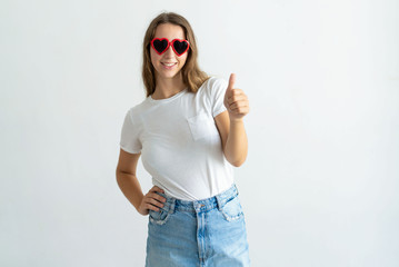 Wall Mural - Woman wearing heart shaped sunglasses and showing thumb up. Smiling pretty young lady looking at camera. Recommendation concept. Isolated front view on white background.