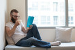 © Mangostar - Concentrated man in glasses drinking coffee while reading book. Serious smart hipster young guy sitting on comfortable sofa. Lifestyle concept