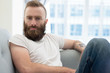 © Mangostar - Pensive bearded guy posing at home. Young man in casual sitting on couch and staring at camera. Leisure at home concept
