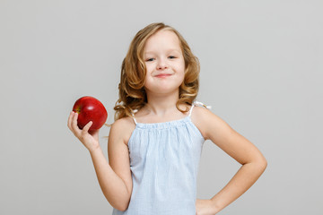 Portrait of a happy smiling little blonde girl on a gray background. Baby eating red apple