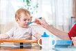 © Artem - Mom feeding the baby holding hand with a spoon of porridge in the kitchen. Emotions of a child while eating healthy food