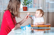© Artem - Mom feeding the baby holding hand with a spoon of food in the kitchen. Healthy baby nutrition. Emotions of a child