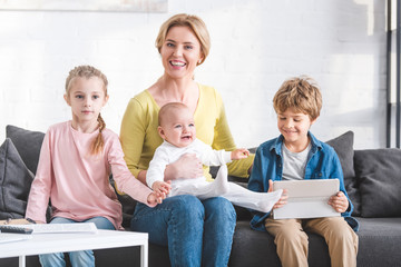 beautiful happy mother with three adorale little kids sitting together on sofa and smiling at camera