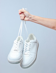 The hand holds by shoelaces hipster white sneakers on gray background. Minimalism.