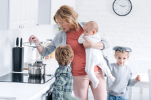 Exhausted Mother Holding Infant Kid And Cooking While Naughty Children Playing In Kitchen