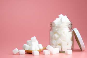 Cube granulated sugar in a glass jar and  on wooden spoon for adding sweetness. some parts was poured on a pink background. - image