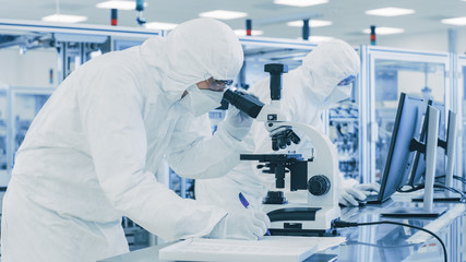 Wall Mural - In Laboratory Scientist in Protective Clothes Doing Research, Using Microscope and Writing Down Data. Workers Working on a Modern Manufactory Producing Semiconductors and Pharmaceutical Items.