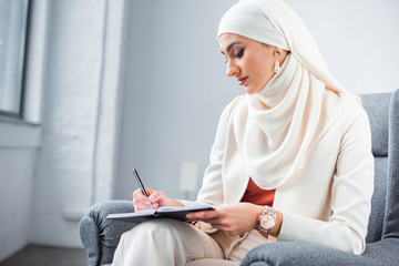 Wall Mural - beautiful young muslim woman sitting in chair and writing in notebook