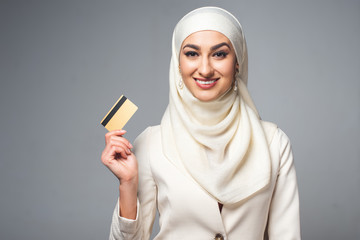 Wall Mural - beautiful muslim woman holding credit card and smiling at camera isolated on grey