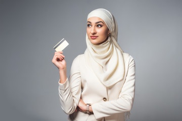 Wall Mural - beautiful smiling muslim woman holding credit card and looking away isolated on grey