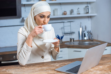 Wall Mural - young muslim woman drinking coffee and using laptop at home