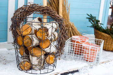 Wicker metal basket with birch wood next to the garden tools at the wall of a blue country house in the snowy winter