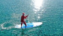 Aerial Photo Of Fit Man Dressed As Santa Claus Practising SUP Or Stand Up Paddle In Exotic Mediterranean Beach With Turquoise Sea