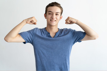 Wall Mural - Positive teenage guy showing body strength. Cheerful young man in casual flexing biceps and smiling at camera. Body strength concept