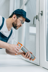 Wall Mural - side view of adult repairman in goggles fixing window with sealant gun