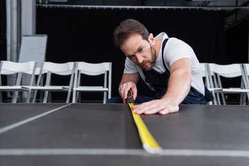 concentrated male craftsman in working overall making measurements of floor by measuring tape