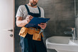 © LIGHTFIELD STUDIOS - partial view of male plumber with toolbelt writing in clipboard near broken sink in bathroom