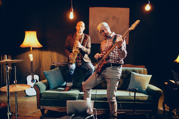 Wall Mural - Young Caucasian man with hat on head playing bass guitar while standing in home studio. In background saxophonist playing his instrument.