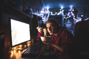 Wall Mural - Portrait of a funny young man eats noodles at night at the computer. Gamer feeds fast food and plays video games at home on the computer. Dinner with a fast noodle soup at the computer.