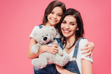 smiling daughter hugging mother and holding soft toy isolated on pink