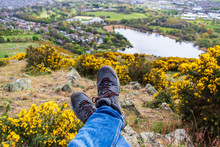 Hiker's Feet With Boots Resting On A Rocky And Green Background With A Distant Lake. Landscape From Arthur's Seat Hill Top In Edinburgh, Scotland. Active Lifestyle Concept.