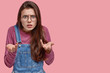 Indignant Caucasian woman looks angrily at camera, gestures with both hands, wears spectacles, looks confused and shocked, models over pink background, blank space for your promotional content