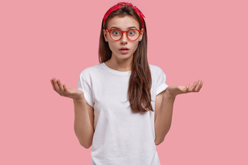 Wall Mural - Horizontal shot of surprised young woman shrugs shoulders, looks in bewilderment, dressed in casual t shirt, wears glasses, looks confusingly, makes decision, models against pink background.