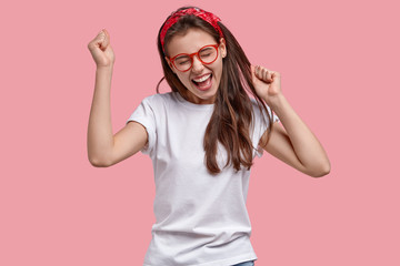 Triumphing joyful woman clenches fists, rejoices positive news, exclaims gladfully, models over pink background, achieves victory and success, isolated over pink background. Overemotive teenage girl