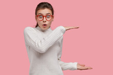 Shocked Caucasian Lady Shows Something Huge, Stares Through Spectacles, Keeps Mouth Opened From Suprise, Dressed Casually Isolated Over Pink Background. People And Size Concept. Look How Big It Is!