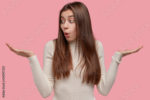 Fotografía Photo of indecisive woman spreads hands, has clueless expression, doubts between two items, wears casual outfit, isolated on pink studio wall, interested in buying new objects