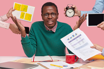 Irritated overworked overload African American guy raises hands with anger, being angry with colleagues who remind him about work, doesnt know from what to start, models over pink studio wall