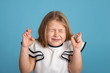 Close up emotional portrait of young blonde  smiling girl wearing white blous with black strips on blue background in studio. She crossed her fingers, shut her eyes and makes a wish