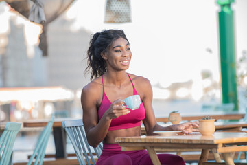 Beautiful tall athletic African American Woman wearing a bright pink workout outfit sits in an outdoor cafe drinking coffee or tea in deep though on a bright sunny day