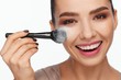 Portrait of a beautiful smiling young woman holding a brush in her hand and applying powder to her face with a brush