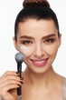© Andriy Goncharenko - Portrait of a beautiful smiling young woman holding a brush in her hand and applying powder to her face with a brush