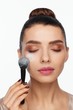 © Andriy Goncharenko - Portrait of a beautiful young woman holding a brush in her hand and applying powder to her face with a brush