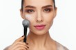 © Andriy Goncharenko - Portrait of a beautiful young woman holding a brush in her hand and applying powder to her face with a brush