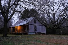 An Old Farmhoue Among The Trees In The Country In The Early Morning Twilight With The Front Porch Light On. Belividere, TN.