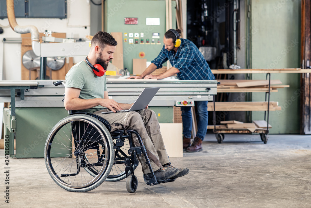 worker in wheelchair with computer in a carpenter's workshop with his colleagu - obrazy, fototapety, plakaty 