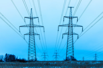 Towers of electric main in the winter snowy countryside field on the background of blue sky and the forest with the wires